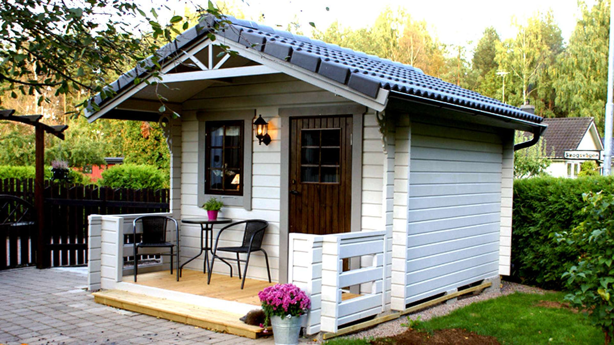 Träfriggebod med verandautbyggnad, vit panel, brunt trästaket och dubbelfönster.